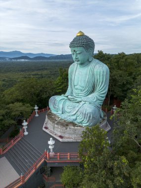 Dev Buda heykeli sakin meditasyon tasvir eder. Heykel, bereketli ormanlarla çevrili, huzurlu ve sakin bir şekilde duruyor. Eski bir bronz figür, ruhsal deneyim sunar.
