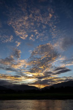 Nefes kesen gün batımı manzarası, parlak turuncu ve altın bulutlarla aydınlatan gökyüzü. Sakin su çarpıcı renkleri yansıtır, görkemli dağ siluetleri üzerinde huzur verici bir manzara yaratır.