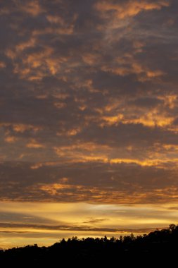 Ateşli turuncu ve altın bulutlarla bezenmiş dramatik günbatımı gökyüzü. Huzurlu bir akşam sahnesi, karanlık manzara siluetinin üzerinde serinlik ve merak uyandırıyor.
