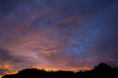 Dramatik günbatımı gökyüzü, sakin doğal manzaranın üzerinde sergileniyor. Renkli bulut oluşumları güzel mor, mavi ve turuncu renkler sergiliyor ve alacakaranlıkta huzurlu bir açık hava manzarası yaratıyor.