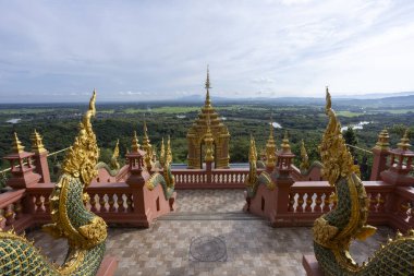 Serene Budist tapınağı mimarisi, altın ve süslü, geniş yeşil manzaranın görkemli panoramik manzarası, uzak dağlar ve geniş bulutlu gökyüzü sunar.