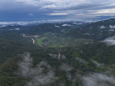 Serene aerial view of mountain landscape with winding river through lush green valley. Dramatic clouds float over dense jungle forest creating peaceful scene