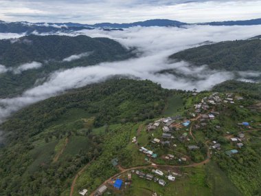 Serene aerial view of remote mountain village in green valley landscape. This peaceful settlement rests on hill above sea of cloud under cloudy sky
