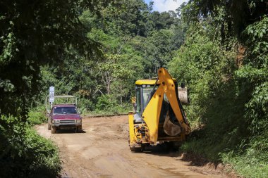 Sarı kepçe yükleyici kırsal ormanda toprak yol inşaatında çalışıyor. Kırmızı pikap, yeşil ormandaki ilerlemeyi ve gelişimi gösteren asfaltsız yolda ilerliyor.