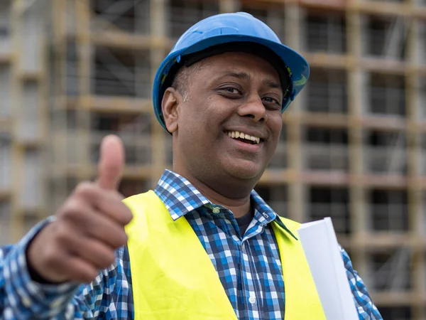 Smiling Indian construction worker wearing safety helmet and vest posing on construction site showing thumbs up gesture