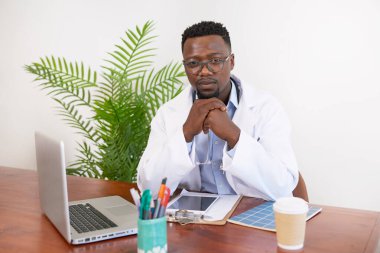Ciddi siyahi doktor ofisinde oturmuş dizüstü bilgisayarla ciddi ciddi bakıyor. Yüksek kalite fotoğraf