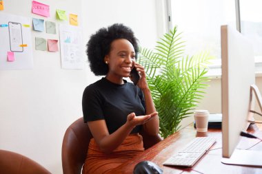 Gülümseyen ve mutlu siyah kadın ofiste telefonla konuşuyor, bilgisayar masasında oturuyor. Yüksek kalite fotoğraf