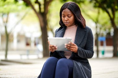Genç siyahi kadın tabletle dışarıda oturuyor, arka planda park ediyor. Yüksek kalite fotoğraf
