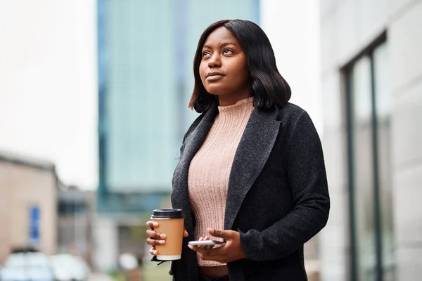 Young Black woman outside office holding cell phone and takeaway coffee. High quality photo