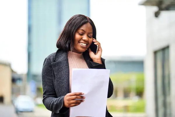 Young Black career woman talking on the phone outside, smiling, holding document. High quality photo