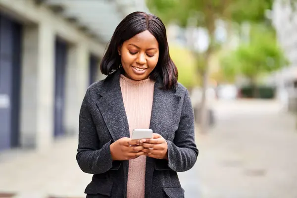 Young Black woman in formal wear looking at cell phone while walking outside. High quality photo