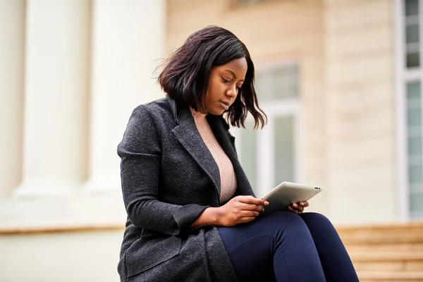 Young Black career woman sitting outside office with mobile device tablet. High quality photo