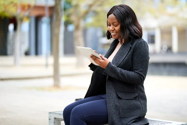 Young Black career woman sitting on outdoor bench with tablet, smiling. High quality photo