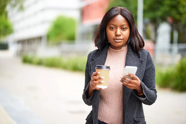 Young Black career woman with cell phone and coffee, business park. High quality photo