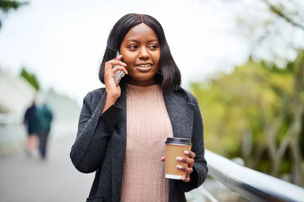 Young Black career woman with takeaway coffee talking on cell phone, urban area. High quality photo