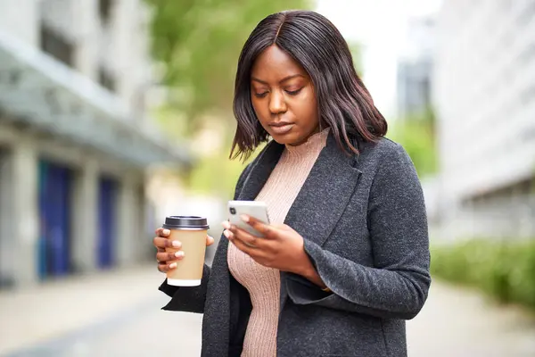 Young Black career woman holding cell phone and takeaway coffee, urban area. High quality photo