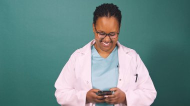 Genç siyahi kadın doktor cep telefonuyla çalışıyor, gülümsüyor ve daktilo ediyor. Yüksek kalite fotoğraf