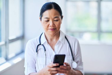 Hastane ortamında akıllı telefon kullanan bir kadın doktor. Tıbbi uzman steteskoplu beyaz bir önlük giyiyor ve sağlık hizmetleri ortamında odaklanma ve konsantrasyon hissi veriyor..
