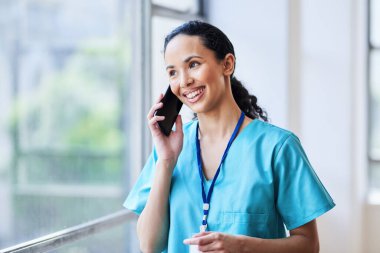 Mavi önlüklü neşeli bir bayan doktor, pencerenin yanında duruyor, bir telefon görüşmesi yapıyor. Görüntü, hastane ortamında profesyonel ve olumlu bir ortam yakalıyor..