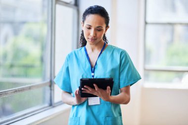 Mavi önlüklü çok ırklı bir kadın doktor, parlak bir hastane ortamında dijital tablet kullanıyor. Görüntü profesyonelliği, sağlık sektöründeki teknolojiyi ve tıp alanındaki çeşitliliği aktarıyor.