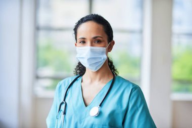 Hastane ortamında yüz maskesi ve steteskop takan kendine güvenen bir doktorun portresi. Tıbbi uzman, parlak ve klinik bir ortamda yeterlilik ve hazırlık yayar..