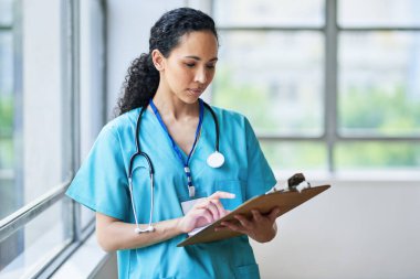 A confident female doctor in scrubs is reviewing patient records on a clipboard in a brightly lit hospital hallway. She represents professional healthcare and dedication to patient care. clipart