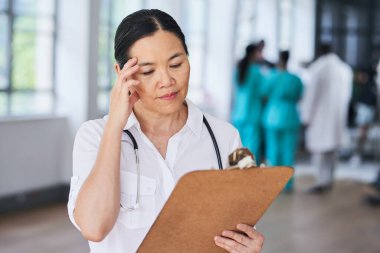 Odaklanmış bir doktor hastane ortamındaki bir panoda hasta bilgilerini inceliyor. Görüntü, arka planda çeşitli sağlık uzmanlarından oluşan bir ekibin bulunduğu yoğun bir tıbbi ortamı gösteriyor..