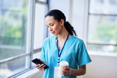 Kadın doktor pencerenin kenarında duruyor, telefonunu kullanıyor ve hastane ortamında kahve fincanı tutuyor. Klinik ortamda sağlık hizmeti, tıbbi profesyonellik ve günlük rutin kavramı.