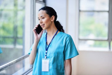 Tıbbi önlüklü bir kadın doktor cep telefonuyla konuşurken, gülümserken ve hastane ortamındaki bir pencerenin yanında konuşurken görülüyor..