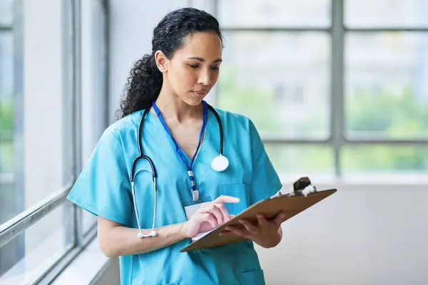 Önlüklü kendine güvenen bir kadın doktor, parlak bir hastane koridorundaki bir panoda hasta kayıtlarını inceliyor. Profesyonel sağlık hizmeti ve hasta bakımına olan bağlılığı temsil ediyor..
