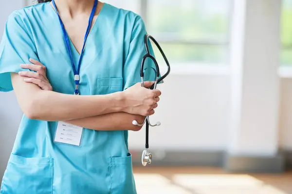 Kolları çapraz duran, steteskop tutan ve hastanede mavi önlük giyen bir doktorun yakın çekimi. Görüntü sağlık hizmetleri, tıp uzmanları ve klinik çevreyi temsil ediyor..