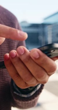 Telefonunu kontrol eden genç bir şehir profesyonelinin yakın çekimi. Adam şehirde duruyor ve gündelik giysiler giyiyor. Modern bir şehir ortamında iş ve bağlantı.