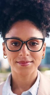 Genç, siyah bir kadın gözlüklü ve büyük bir gülümseme ile şehir iş ortamında. Kıvırcık saçları var ve spor ceket giyiyor. Ona profesyonel bir görünüm veriyor. Şehir ortamı, finans bölgesi.