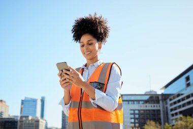 Modern şehir şantiyesinde akıllı telefon kullanarak yüksek görüşlü yelek giyen gülümseyen kadın inşaat işçisi. Mutlu ve kendinden emin, inşaattaki teknolojiyi kucaklayan. Modern şehir manzarası