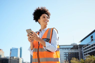Kentsel çevrede kendine güvenen kadın inşaat işçisi, akıllı telefon tutuyor ve arka planda modern binalar bulunan bir güvenlik yeleği giyiyor..