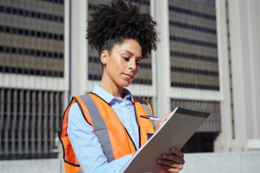 Güvenlik yeleği giymiş genç bir kadın mühendis elinde bir pano, bir inşaat alanını inceliyor, şehir merkezinde..