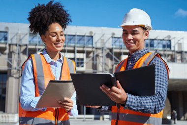 Güneşli bir günde bir şantiyede proje planlarını tartışan güvenlik yelekleri ve sert şapkalı iki farklı inşaat işçisi..