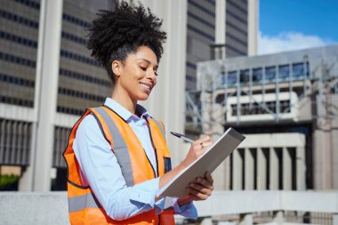 Kadın inşaat işçisi, güvenlik yeleği giyiyor, gülümsüyor ve şehir binasının dışındaki bir panoya not alıyor..