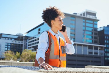 Güvenli yelek giyen kendine güvenen kadın inşaat işçisi arka planda modern şehir binaları olan açık havada telefon kullanıyor. Profesyonelliği, iletişimi ve şehir hayatını temsil ediyor.