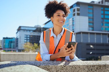 Turuncu güvenlik yeleği giymiş gülümseyen kadın inşaat işçisi dışarıda mola veriyor, elinde akıllı bir telefon, arka planda şehir binaları var..