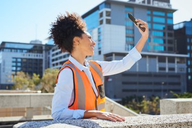 Turuncu yelek giyen bir kadın inşaat işçisi, şehir ortamında gülümseyen akıllı telefonuyla selfie çekiyor..