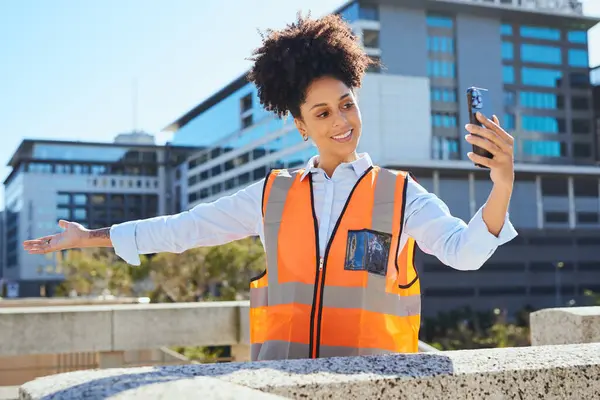 Güneşli bir günde şantiyede selfie çeken parlak güvenlik yeleği giymiş kadın inşaat işçisi. Arka planda şehir binaları.