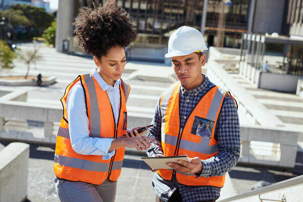 Two diverse construction workers in safety vests and hard hats reviewing plans on a city project outdoors.