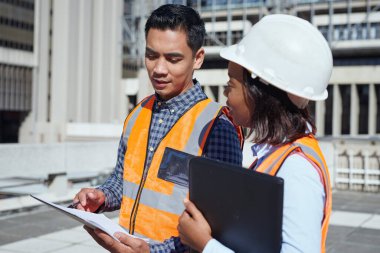 İki farklı mühendis bir inşaat sahasında plan üzerinde çalışıyor. Biri güvenlik kaskı takıyor, ikisi de koruyucu yelek giyiyor..