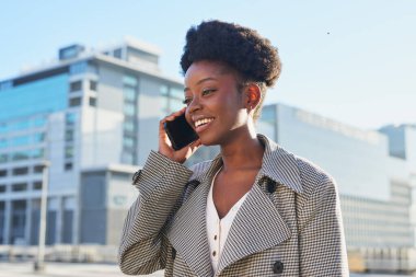 Neşeli bir yüzü olan genç bir kadın şehir ortamında akıllı telefonuyla konuşuyor. Şehirde güneşli bir günün tadını çıkarırken kendine güveni ve mutluluğu yayar..