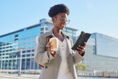 Kendine güvenen bir iş kadını modern bir ofis binasının önünde duruyor, sabah kahvesinin tadını çıkarıyor ve tablet kullanıyor.. 