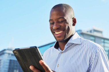 Neşeli bir adam şehir merkezinde dışarıda dururken bir tablet kullanıyor. Parlak, güneşli bir gün, olumlu ve üretkenlik hissini artırır..