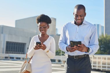 İki profesyonel, mobil cihazlarıyla aktif olarak meşgul olan bir şehir ortamında görülüyor. Görüntü, modern bir şehir ortamında üretkenlik ve bağlantı hissini yakalıyor..