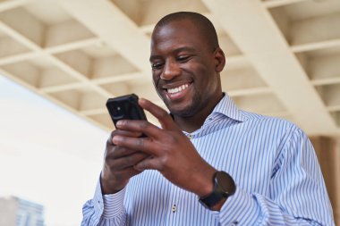 Çizgili gömlek giyen neşeli bir adam modern mimari ortamlarda akıllı telefonunu kullanmaktan hoşlanır. Görüntü, bir bağlantı ve olumlu duyguyu yakalar.