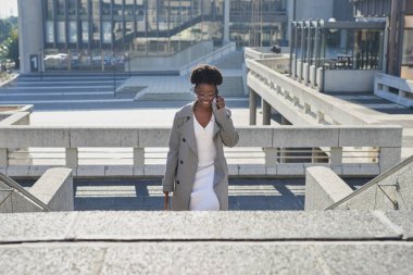 Profesyonel bir kadın modern bir şehir ortamında güvenli bir şekilde beton basamaklara tırmanıyor. Bir telefon görüşmesi yapıyor, şık gözlükler takıyor ve şık bir ceket giyiyor.
